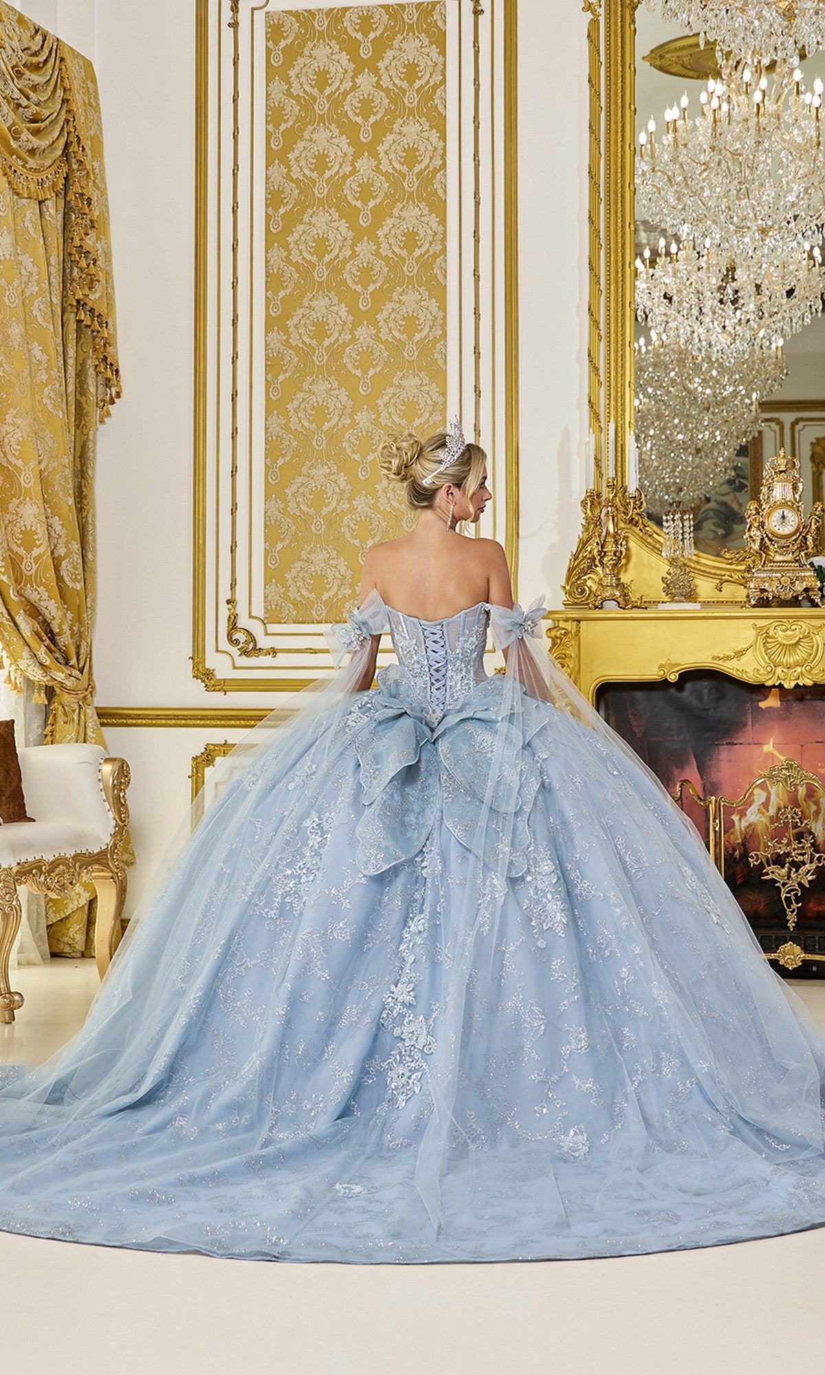 A woman wearing the Quinceanera Dress 5074 By Dancing Queen, a light blue ball gown with a sheer corset bodice, stands with her back to the camera in an opulent gold room with a grand chandelier and elegant fireplace.
